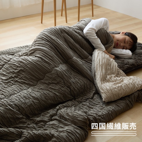 一名年輕女子穿著白色長袖上衣，正舒適地側躺在地板上的深灰色鋪棉寢具組中，身上蓋著同樣具有皺褶紋理感的深灰色被子，露出內層淡米色的設計，畫面右下角帶有四國纖維販賣的品牌文字。