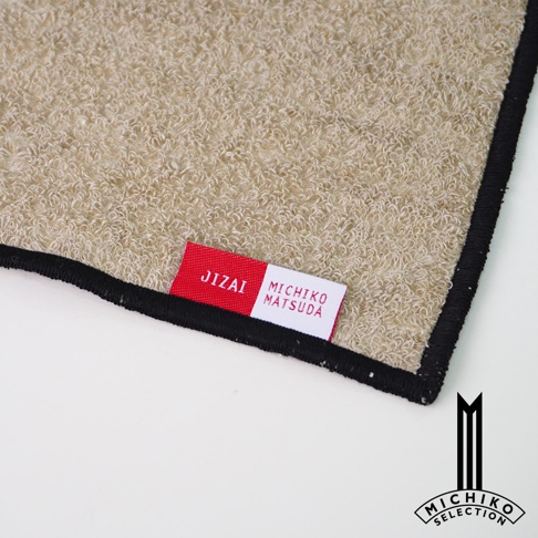 一張米色觸感毛巾的特寫，角落有紅色和白色標籤，上面印有「JIZAI」和「MICHIKO MATSUDA」字樣，下方是「MICHIKO SELECTION」標誌。