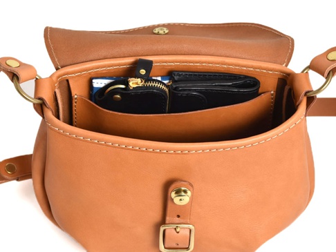 職人手作皮革休閒旅行馬鞍包，淺棕色牛皮材質，包身縫線清晰。包內有一個黑色拉鍊零錢包和一個藍白條紋卡夾。包蓋上飾有金色金屬釦。