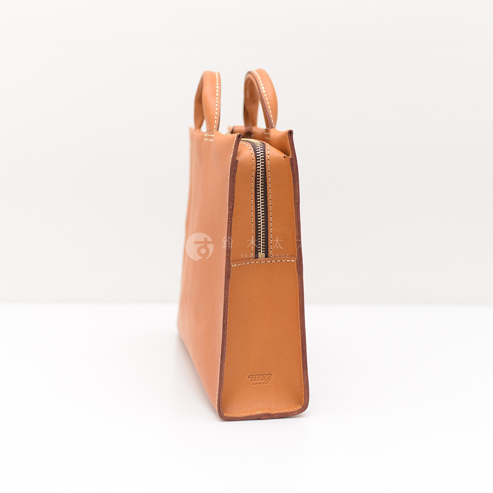 日本 HERZ 職人手作皮革簡約商務公事包，展示包包的側面，皮革材質呈現溫潤光澤，簡約設計風格，頂部有提把，側面有拉鍊，底部壓印有 HERZ 品牌標誌