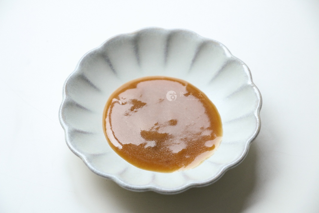 淺灰色花瓣形狀的陶瓷小碟中，盛有濃稠的棕褐色芝麻沾醬，醬料表面光滑，帶有細微光澤。