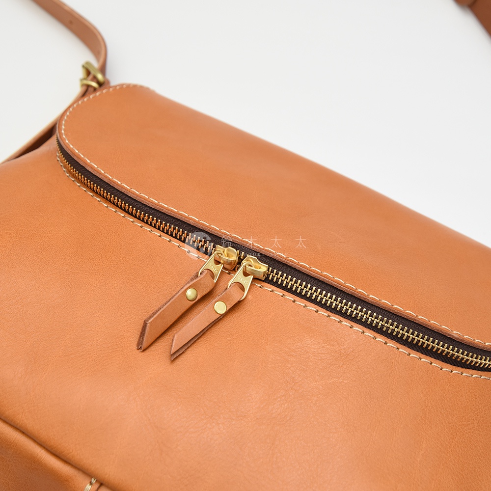 以駝色皮革製作的斜背包，特寫鏡頭聚焦在兩個金色的雙拉鍊頭和皮製拉繩，拉鍊旁有精緻的縫線細節，呈現職人手作的質感。