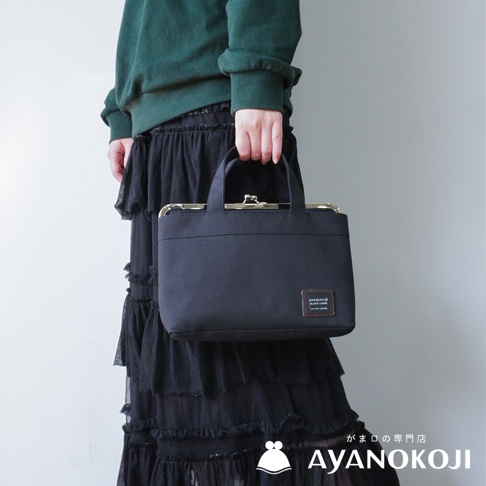 一位身穿深綠色毛衣和黑色荷葉邊長裙的女子，手提一個黑色橫式口金手提包，包上印有AYANOKOJI的品牌標籤。