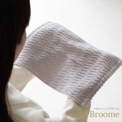 Broome 今治柔韻波紋手帕，灰白色調，表面有細緻的波浪緹花紋理，由一位女性手持，背景為柔和的白色光線，右下角有 Broome 品牌標誌。