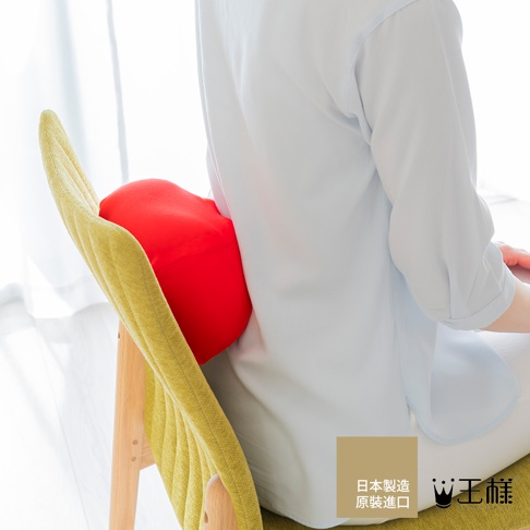 一個紅色的指壓按摩枕，放置於椅背上，由一位穿著淺藍色上衣和白色褲子的人使用。按摩枕採用柔軟的布料材質，形狀類似心臟，提供腰部支撐。