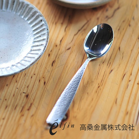 日製錘目紋不鏽鋼餐匙，銀色，勺面光滑，勺柄帶有錘擊紋理，放置於木紋桌面及白色紋理餐盤上。