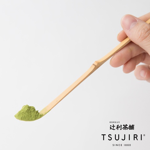 一根天然竹製的抹茶杓，末端盛著一小撮翠綠色的抹茶粉，由一隻手持握，背景為純白色。