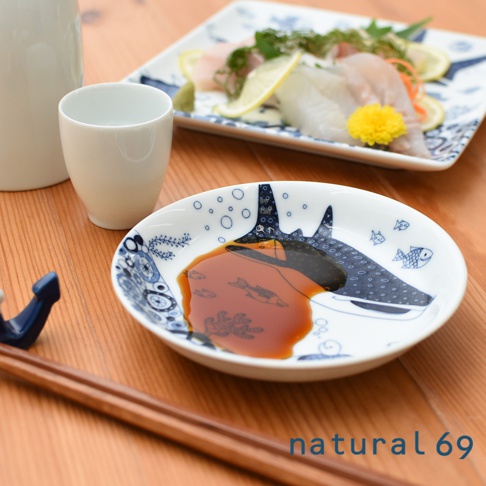 帶有藍色海洋生物圖案的白色陶瓷小碟，盛有醬油，旁邊是白色陶瓷杯和木質筷子，後方盤子裡有生魚片。