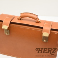 簡約的淺棕色皮革機師箱，配有金色金屬鎖扣、皮質提把與縫線細節。箱身為光滑皮革材質，質感細緻。