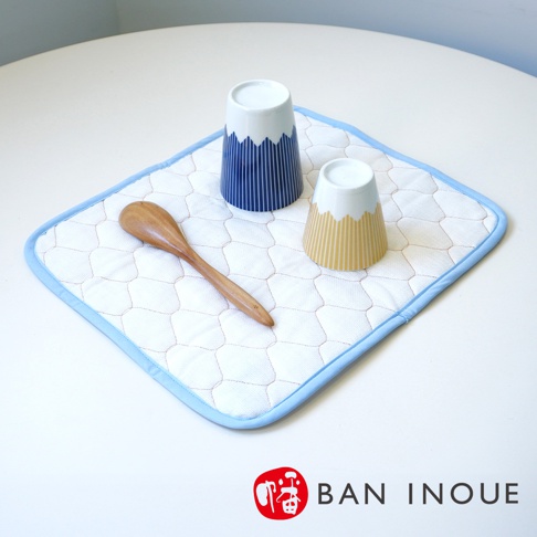 白色棉織吸水墊，邊緣為淺藍色滾邊，墊上飾有淺紅色網狀縫線。墊上擺放著一個木製湯匙，以及兩個富士山造型的杯子，一個為藍色條紋，另一個為黃色條紋。