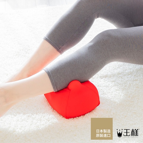 一個紅色的指壓按摩枕，形狀類似心臟，放置於鋪有白色絨毛地毯的地面上，一名穿著灰色七分褲的女性雙腿彎曲，將腳踝置於按摩枕上。