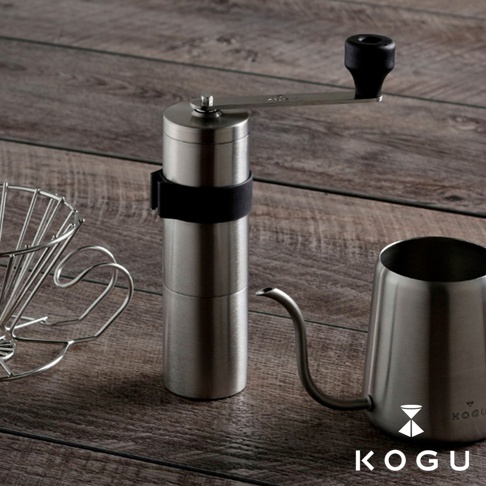 銀色18-8不鏽鋼手搖咖啡磨豆器，配有黑色手柄及黑色固定帶，置於木紋背景上，旁邊有同色系的金屬濾網架和細嘴咖啡壺。