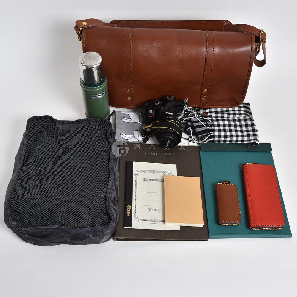 職人手作皮革復古郵差包，棕色皮革，配有金屬釦帶。包內裝有一台相機、綠色保溫瓶、黑色網格收納袋、黑白格紋布、筆記本和卡夾，展現多功能收納。