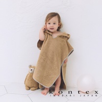 一名嬰兒穿著棕色連帽浴巾，浴巾帶有可愛的熊耳朵設計。浴巾材質柔軟，邊緣為灰色滾邊。一旁擺放著一隻泰迪熊玩偶和一個白色氣球。