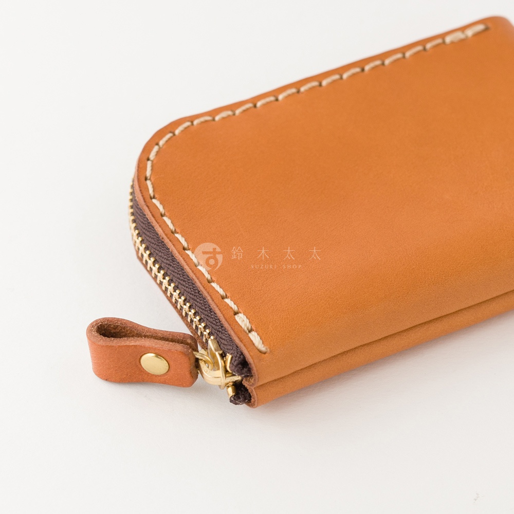 一個淺棕色職人手作皮革零錢包的特寫視角，邊緣採用粗縫線設計，深色拉鍊搭配金色拉鍊頭與皮質拉環，包身中央印有鈴木太太的品牌標誌。