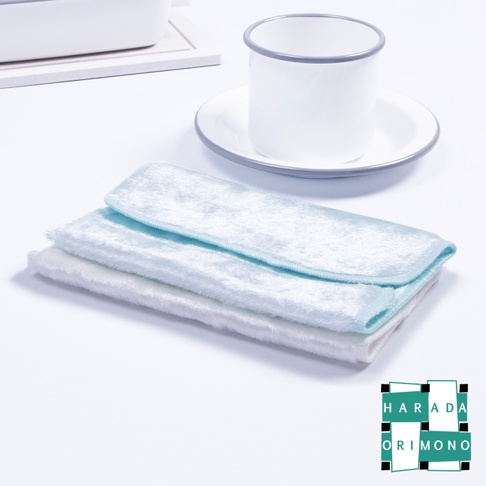 淺藍色與白色兩塊絨毛材質的廚房專用布疊放，布料柔軟蓬鬆，邊緣帶有淺藍色滾邊。背景為白色桌面，左側有白色方形盤，右側有白色馬克杯與白色盤子，杯子與盤子邊緣有灰色細線。
