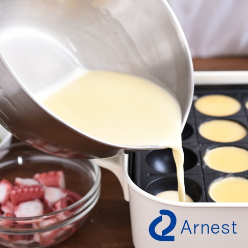 Arnest 品牌的不鏽鋼調理碗正傾斜著倒出金黃色麵糊，倒入熱騰騰的章魚燒烤盤中，旁邊還放著一碗切塊的章魚。