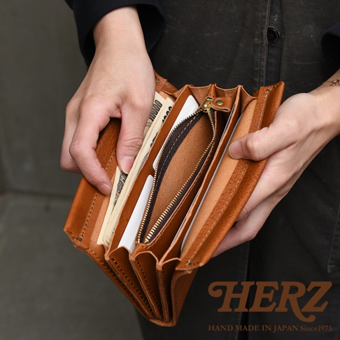雙手拿著日本HERZ品牌職人手作皮革長夾，呈現皮夾開啟後的內部結構，皮革呈現溫潤的棕褐色澤，質感厚實且紋理自然，中間設有拉鍊零錢袋，兩側分層放置紙鈔與卡片，背景為素色空間，右下角印有HERZ品牌名稱與相關標語。