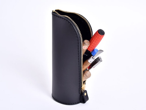 黑色皮革製直立式筆袋，頂部拉鍊開口，內含多支筆，其中一支為紅色格紋握把。