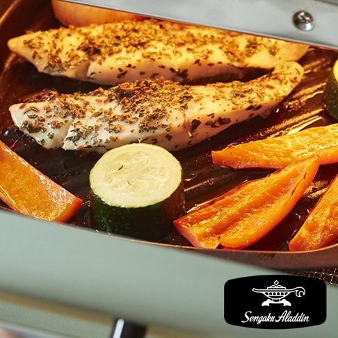 烤箱內有兩塊香草烤魚，旁邊擺放著切片的櫛瓜、紅甜椒和南瓜。烤盤是黑色的，烤箱外殼呈淺綠色。