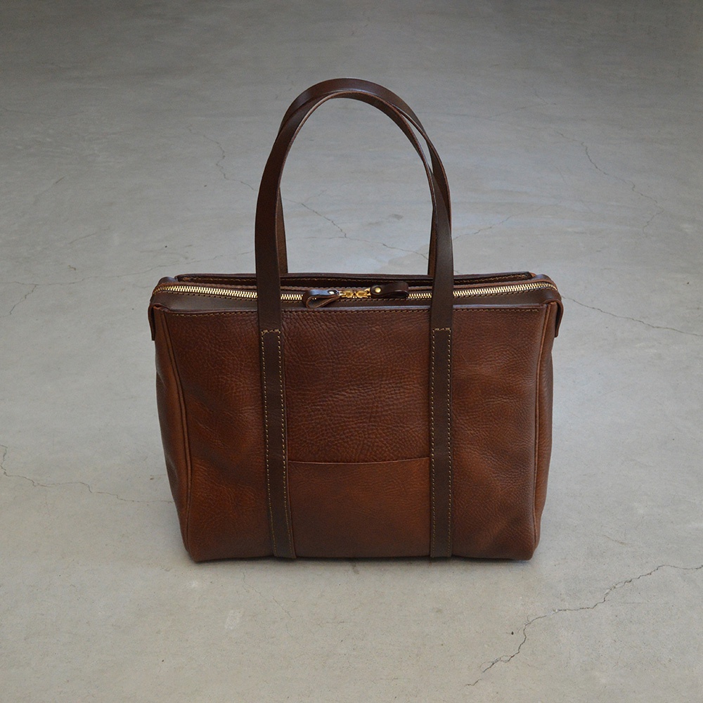 棕色皮革手提肩背包，附有拉鍊開口和兩個提把，包包正面有一個方形口袋，皮革表面有自然紋理。