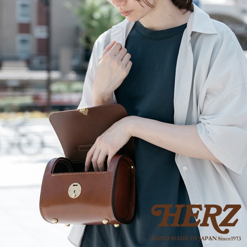 職人手作皮革復古杜勒斯手提包，採用棕色皮革製作，包身呈現圓筒狀，配有金色鎖扣和底部防磨釘。