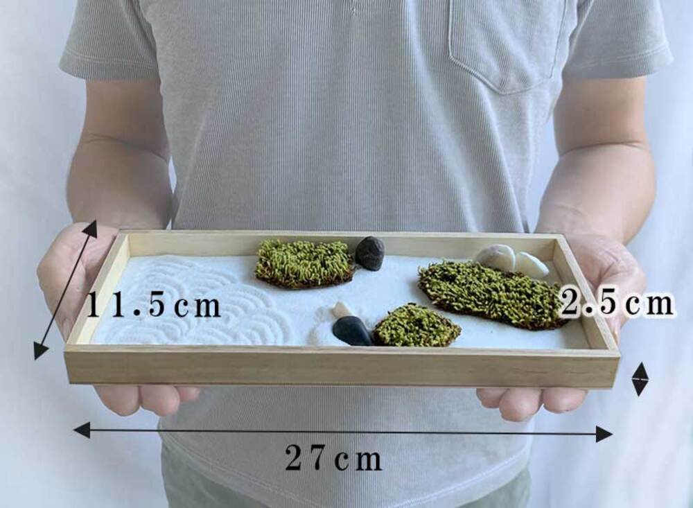 一個淺木框，內含白色細砂，砂上耙出波紋，點綴著幾塊綠色苔蘚裝飾和深淺不一的黑色、灰色及白色鵝卵石。