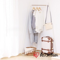 白色角落造型吊掛衣桿，上掛有灰色長袖外套、條紋長袖上衣和白色長袖上衣，旁邊掛著米白色斜背包，下方有竹編籃和復古風格行李箱。