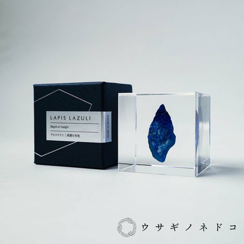 一個透明立方體內嵌著一顆深藍色、帶有金色斑點的青金石礦石，旁邊是一個深藍色的盒子，盒蓋上印有「LAPIS LAZULI」及日文「ラピスラズリ | 深淵な知性」。