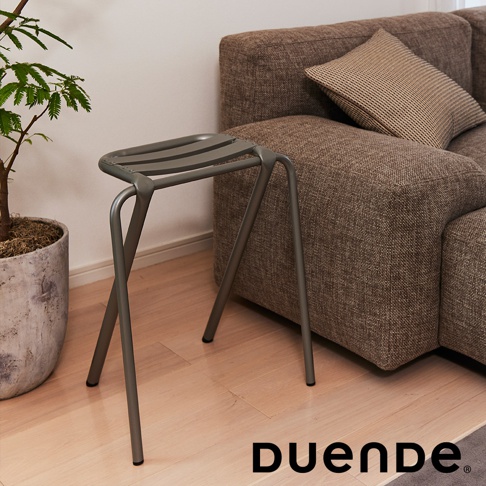 灰色金屬材質的吧檯椅，椅面為條狀簍空設計，椅腳為細長的金屬管組成，與後方的棕色布紋沙發和盆栽一同置於木地板上。