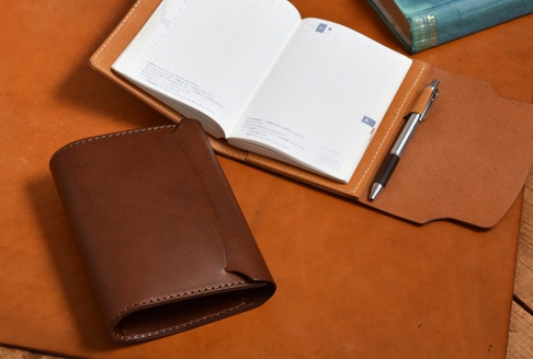 職人手作皮革A6記事本書套，深棕色牛皮材質，搭配一本打開的日誌本和一支銀色筆，整體陳列於淺棕色皮革桌面。