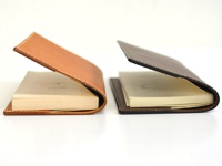 兩款職人手作皮革厚革書套，一款為淺棕色牛皮，另一款為深棕色牛皮，均搭配白色車縫線。書套內含薄頁書籍，呈現簡約質感。