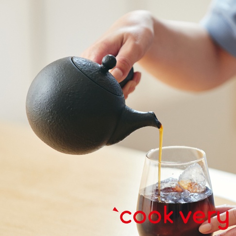 一只手正傾斜著一個黑色、帶有粗糙紋理的常滑燒咖啡急須茶壺，壺嘴流出咖啡液，倒入裝有冰塊的玻璃杯中。