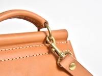淺棕色皮革迷你後背包的頂部提把和肩帶扣環特寫。皮革表面有細緻的縫線，金屬扣環為黃銅色。