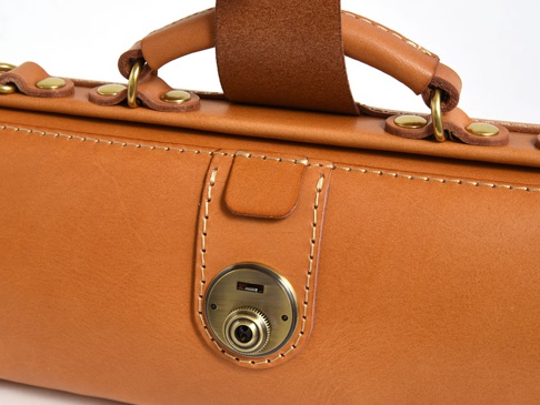 職人手作皮革橫長形杜勒斯單肩包，採用天然棕色皮革，配有黃銅色金屬鎖扣和铆釘，以及皮質提把和肩帶固定環。手工縫線清晰可見，展現精緻工藝。