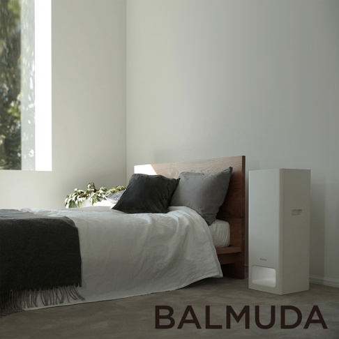 The Pure 空氣清淨機，白色直立式機身，簡約設計，放置於木製床架旁，搭配白色床單、灰色枕頭與深灰色毛毯，窗外綠意透入。