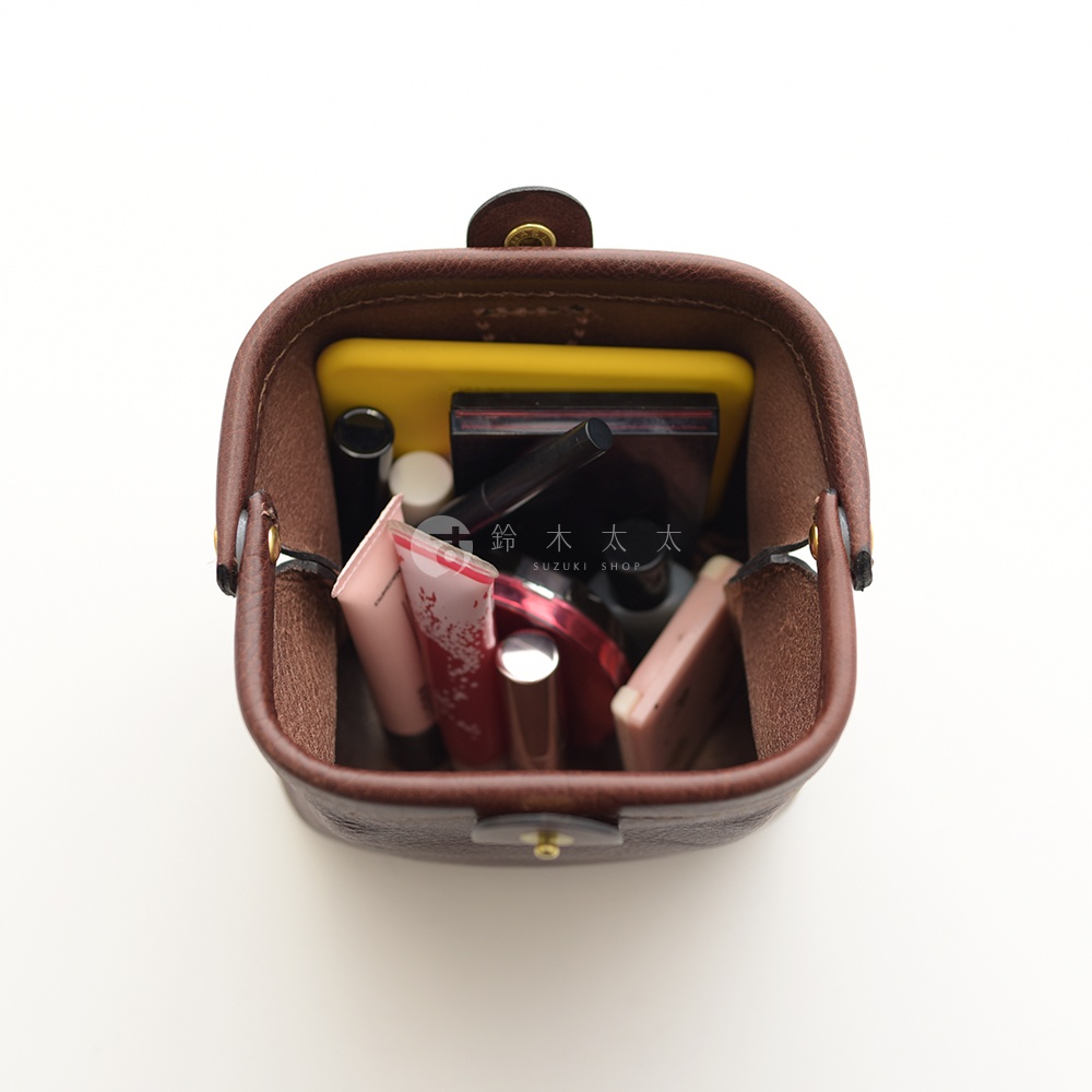 深棕色皮革製作的職人手作口金化妝包，內部裝有手機、唇膏、眼線筆、粉餅等化妝品。