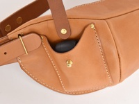 職人手作皮革斜背包，呈現天然原色，材質為真皮。包款設有外側小口袋，可放入耳機等小物，並以銅質釦件與縫線裝飾，展現手工質感。