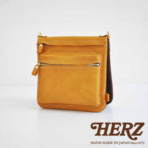 職人手作皮革翻轉雙色直式斜背包，主體為芥末黃色，搭配深棕色內襯，採用翻蓋式設計，正面有一個拉鍊口袋，整體為直式斜背包造型。