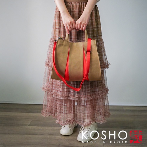 一位女性手提著一個淺棕色棉帆布扇形托特包，包上有兩條鮮紅色裝飾條和背帶。