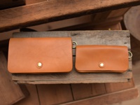 兩個棕褐色皮革長夾，一個較大，一個較小，皆配有金色按扣和側邊的金色圓環，擺放在木質表面上。