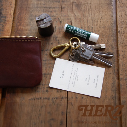 酒紅色職人手作皮革Organ拉鍊式小物收納袋，搭配金色金屬拉鍊。旁邊有鑰匙圈、幾串鑰匙、一條護唇膏，以及一張印有品牌資訊的白色小卡。