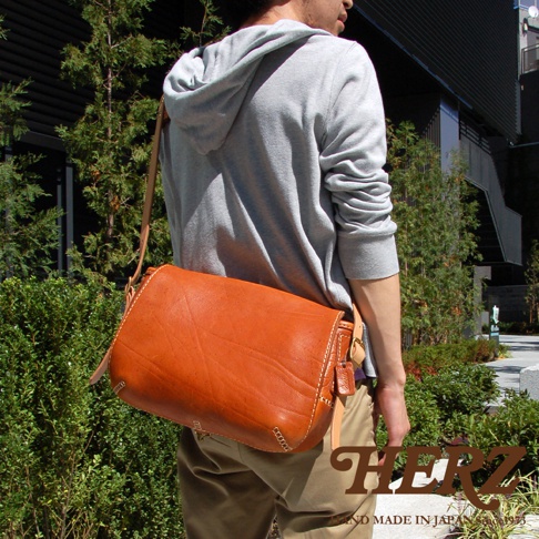 男性斜背著一個橘棕色皮革郵差包，包包有明顯的縫線和皮革紋理，包蓋上有一個扣環。