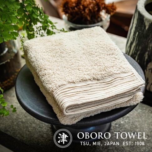 一條米白色、觸感蓬鬆的毛巾，帶有羅紋邊飾，放置於黑色圓形底座上。