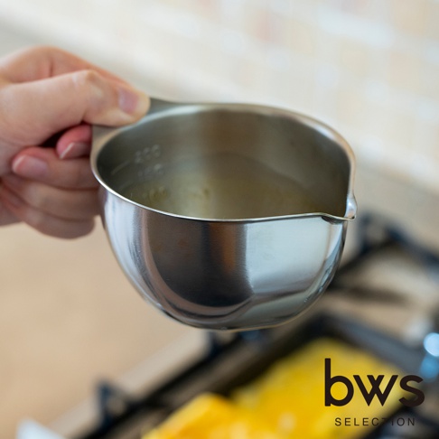 一隻手拿著一個 bws SELECTION 品牌的不鏽鋼料理備料盆，杯緣處有刻度標示，備料盆前方有傾倒用的壺嘴設計，下方背景模糊處可見正在烹飪的黃色蛋液食材。