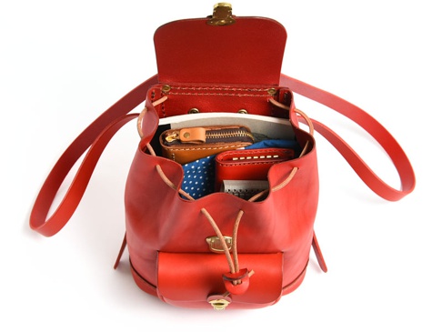 鮮紅色皮革迷你後背包，包身開口處有抽繩設計，內部裝有皮夾、零錢包與藍底白點布料。包身正面有一個翻蓋小口袋，配有束帶與金屬鎖扣。