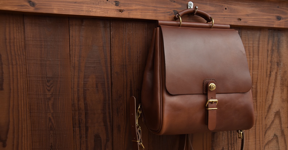 一個棕褐色的皮革後背包，具有屋瓦狀的翻蓋設計，搭配金色扣環和金屬配件，懸掛在木質背景上。