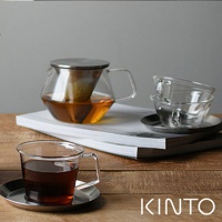 Carat 茶壺，容量 600ml，由透明玻璃製成，帶有金屬濾網和把手，壺內盛有琥珀色茶飲。旁邊還有兩個玻璃杯和一個茶杯，皆放在銀色茶碟上。