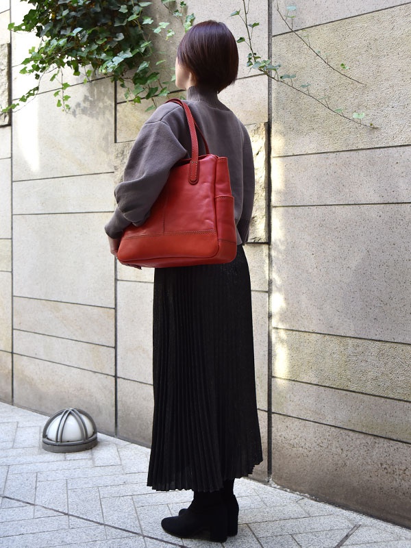 一位女性身穿深灰色高領毛衣與黑色百褶長裙，肩背一個紅色皮革直式托特包，包包有兩個側袋，提把與包身相同材質。