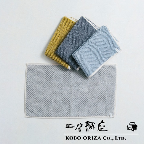 三塊chevron紋理的毛巾，分別為芥末黃、深灰藍和淺藍色，疊放在一同色系的灰色chevron紋理浴墊上方。